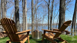 Dos sillas Adirondack en un área con césped, un brasero metálico y vistas a un lago y árboles.