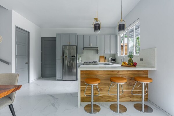 A modern kitchen with grey cabinets, a stainless steel refrigerator, a wooden island with bar stools, and pendant lighting.