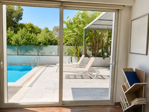 A room with a sliding glass door leading to a pool area with lounge chairs and a pergola. - Welcome to Lumen Villa the First of the Adelos Trilogy (Elliniko-Argyroupoli)