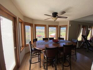 A dining area with a round table, six chairs, a ceiling fan, and large windows offering a view of a rural landscape.