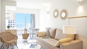 A modern living room with a sofa, a coffee table, a wicker chair, and a dining area with a table and chairs. There are two wall-mounted mirrors and a lamp on a stand. - BJ Cala Morlanda Estudios (Manacor)