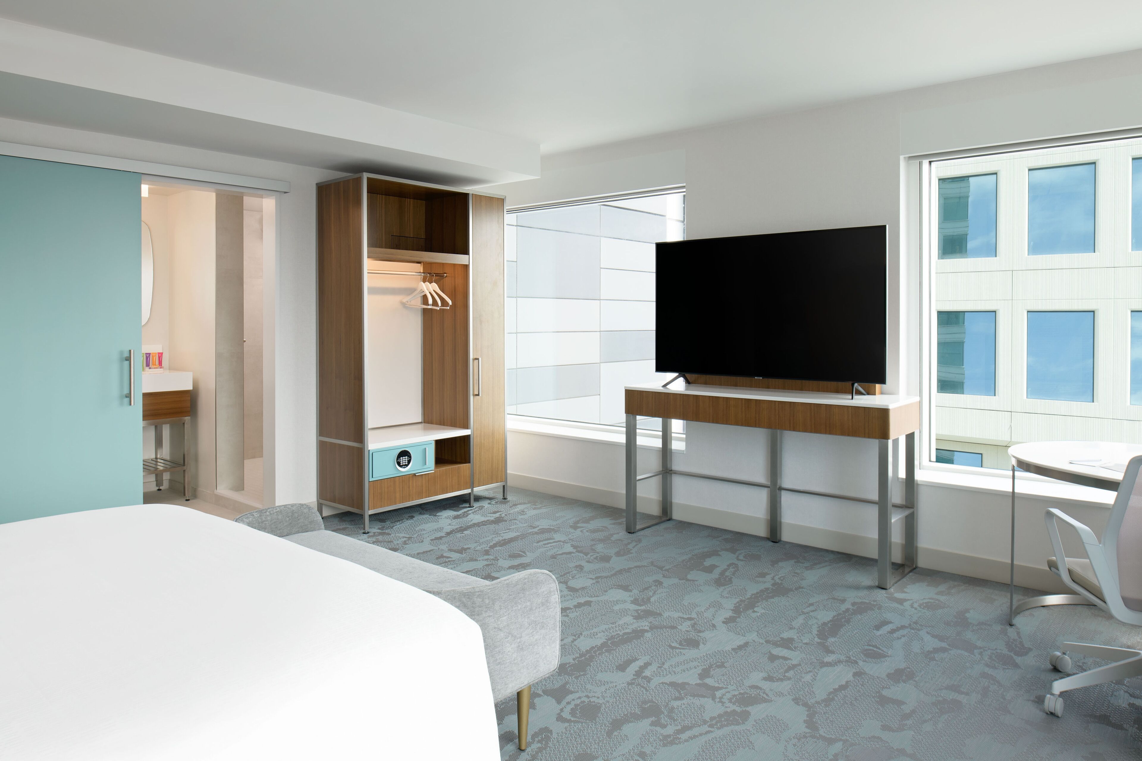 A modern hotel room with a bed, a desk with a flat-screen TV, a wardrobe, and a window with a city view.