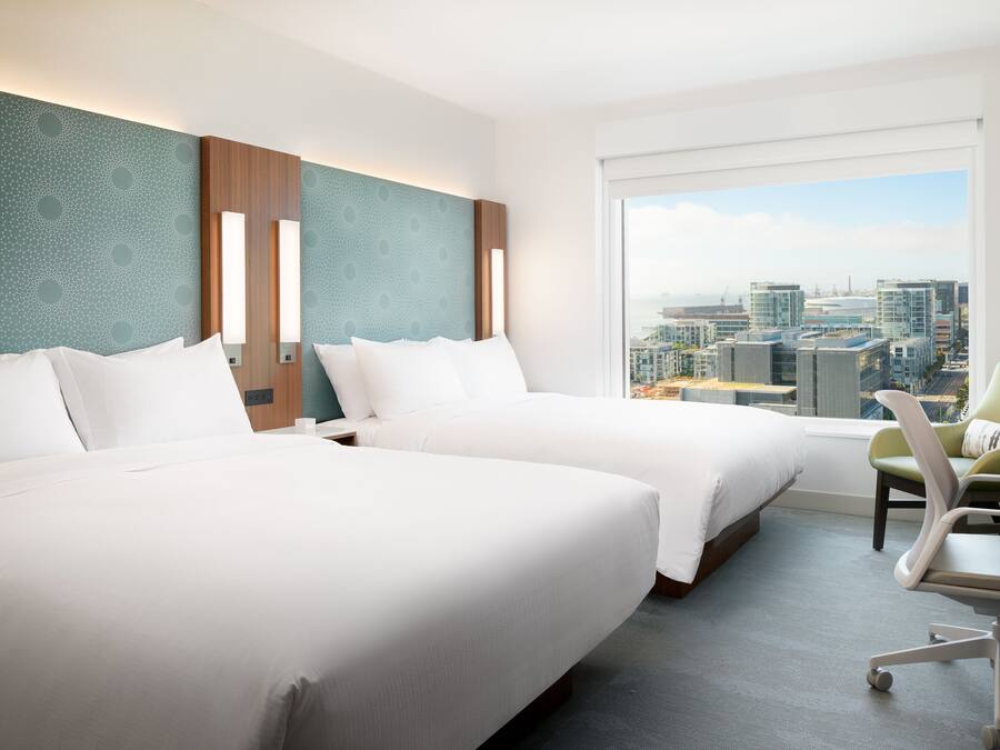 A hotel room with two beds, a desk, and a view of a cityscape.