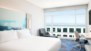 Ein Hotelzimmer mit einem großen Bett, einem Schreibtisch, einem Stuhl und Blick auf die Stadt durch große Fenster.