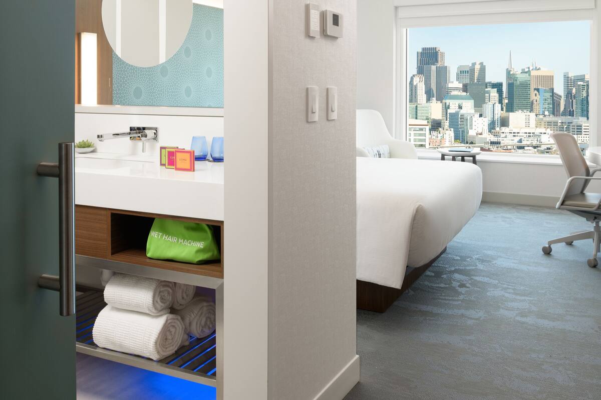 Hotelkamer met uitzicht op de stad, een bed, een bureau en een badkamer met spiegel en wastafel.
