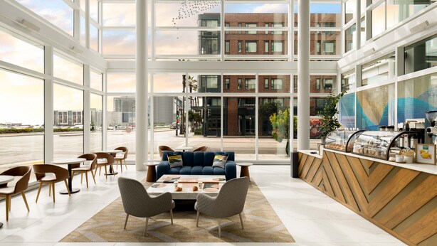 Eine großzügige Lobby mit großen Fenstern, einer Sitzecke mit einer Couch und Sesseln sowie einem Empfangstresen mit einer Kaffeestation.