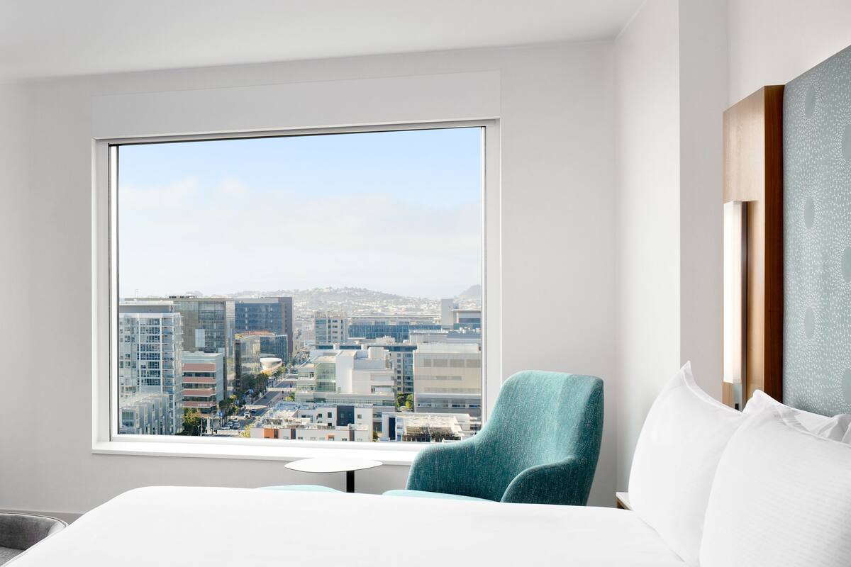 Een hotelkamer met een groot bed, een smaragdgroene fauteuil en een stadszicht door een groot raam.