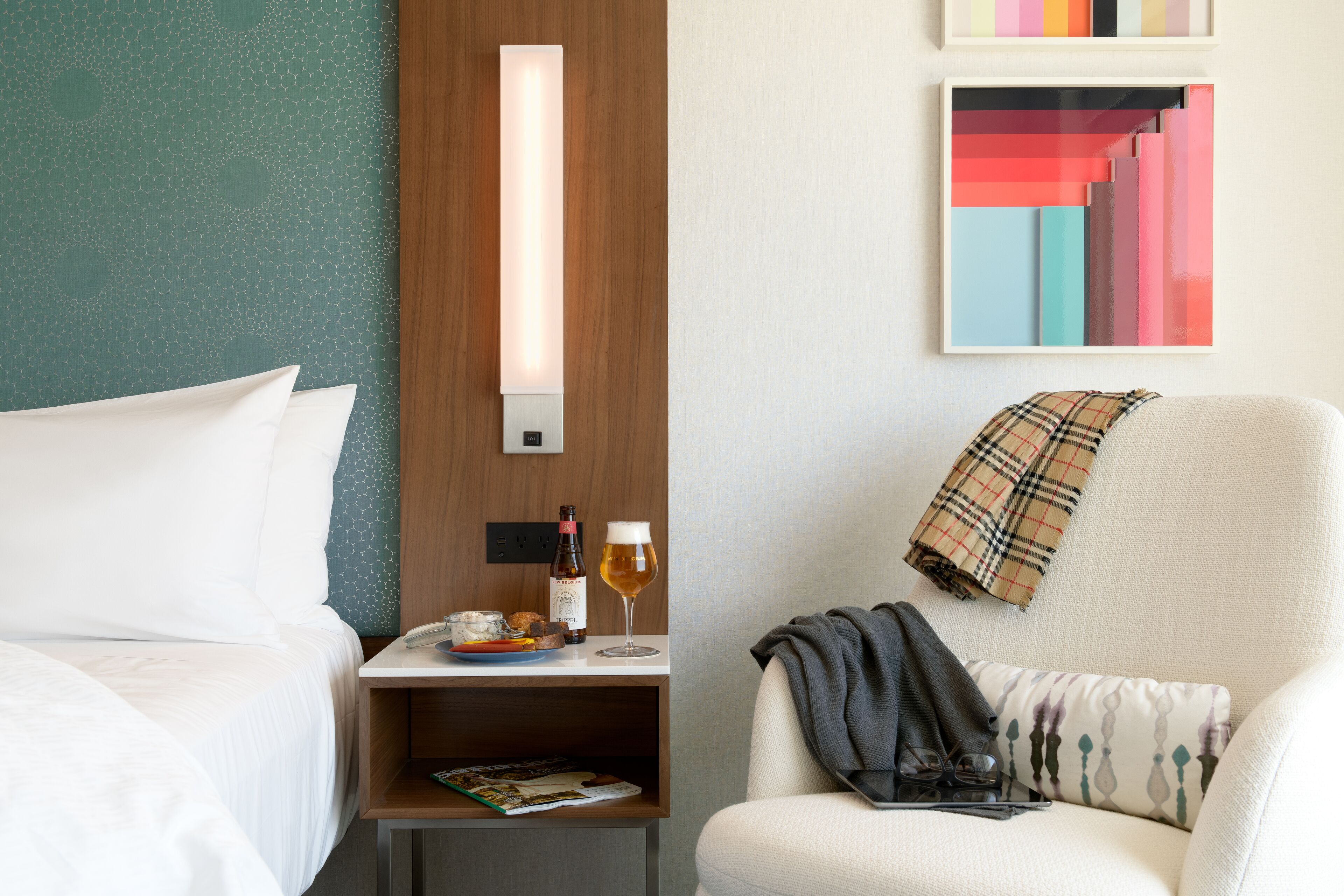 A hotel room with a bed, a nightstand with a bottle and glass, a chair with a blanket, and a wall with a geometric painting.