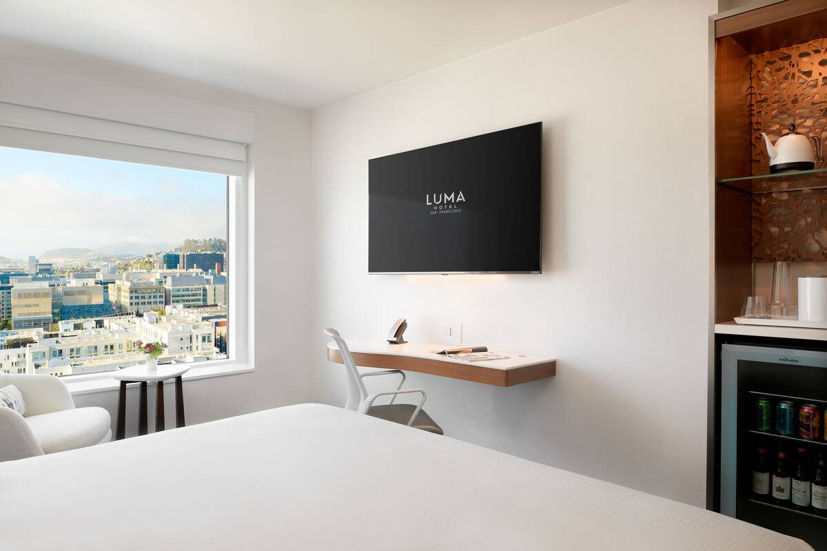 Een moderne hotelkamer met een groot raam dat uitzicht biedt op de stad, een flatscreen televisie aan de muur, een klein bureau met een stoel, en een minibar met drankjes.