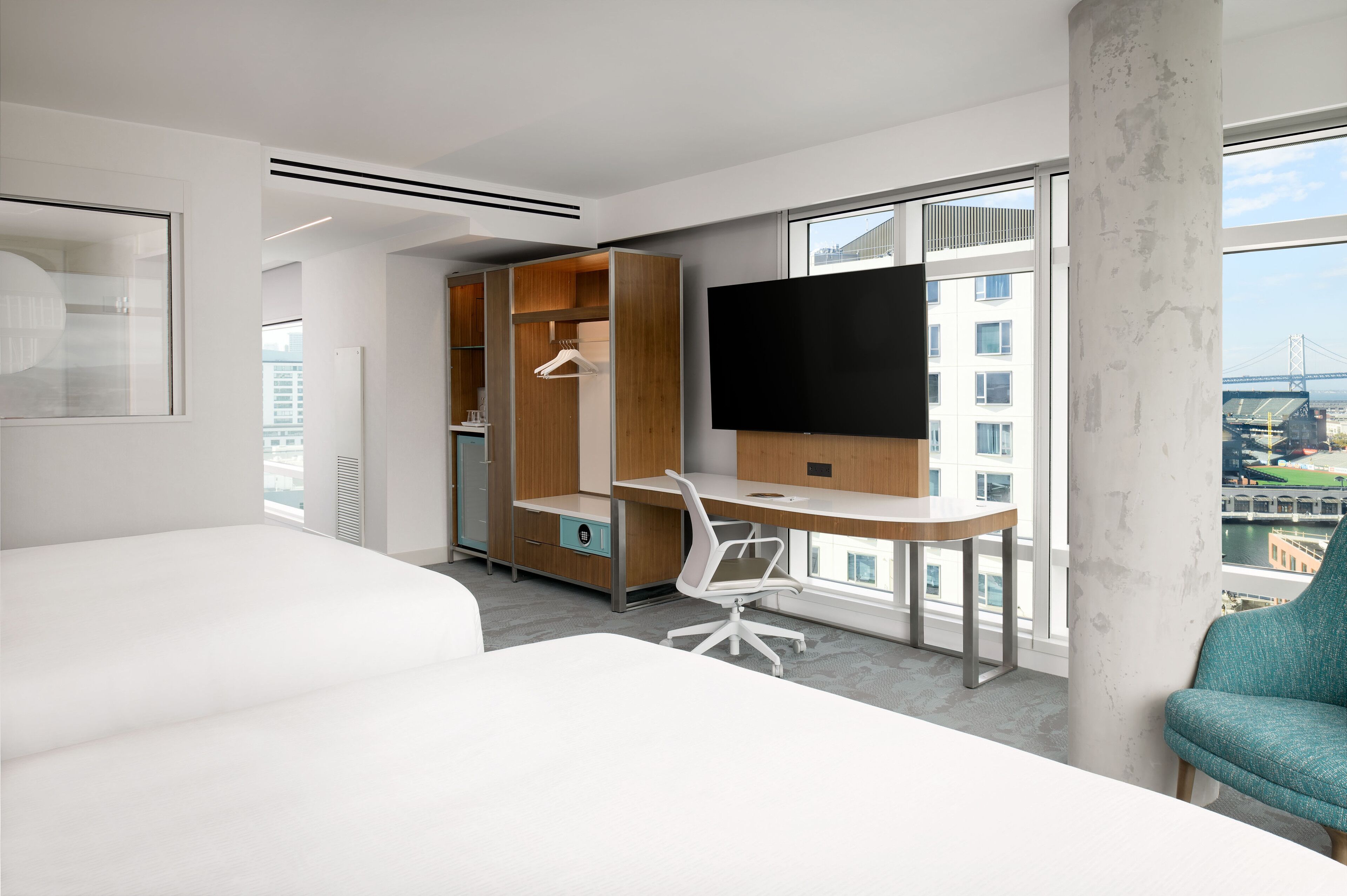 A modern hotel room with a large bed, a desk with a chair, a wardrobe, a TV, and a view of a cityscape through large windows.