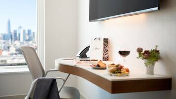 A modern hotel room with a desk, a chair, a flat-screen TV mounted on the wall, a vase with flowers, and a view of a cityscape through the window.