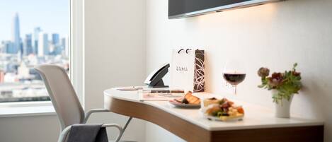 A modern hotel room with a desk, a chair, a flat-screen TV mounted on the wall, a vase with flowers, and a view of a cityscape through the window.