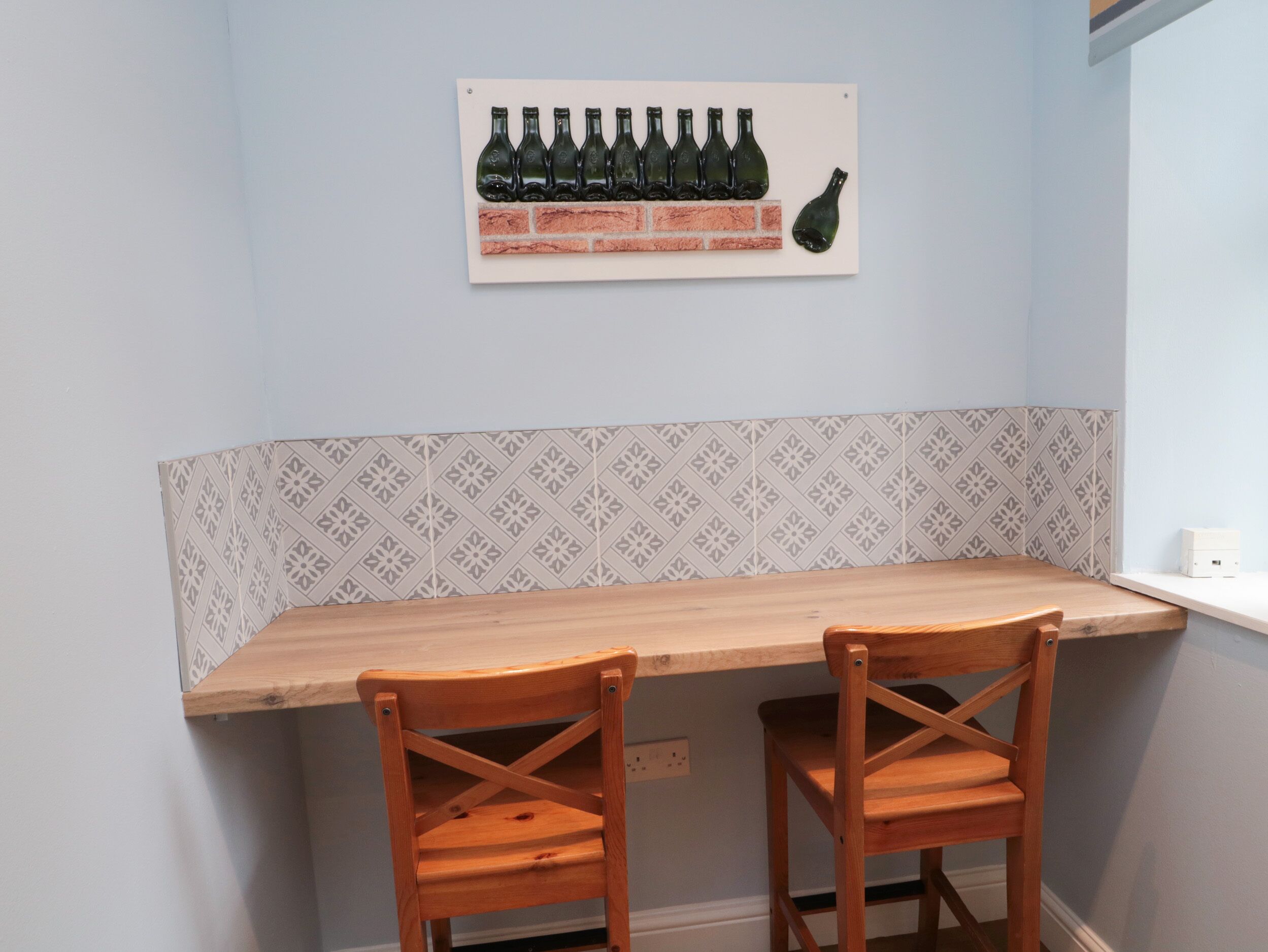 A corner kitchen with a wooden table and two chairs, a backsplash with a geometric pattern, and a wall-mounted art piece of bottles.