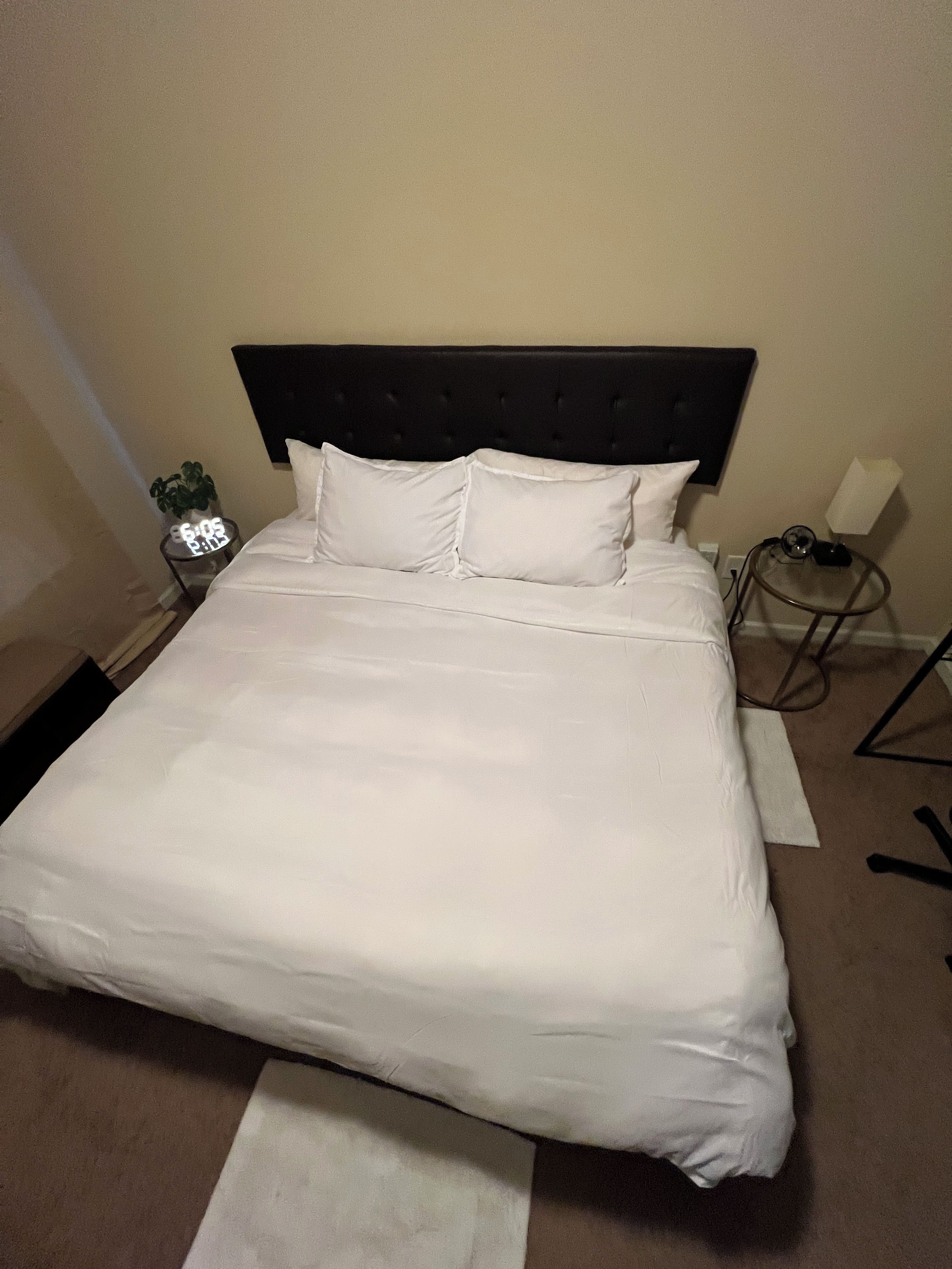 A neatly made bed with white linens, a dark headboard, and a bedside table with a lamp and a small plant.