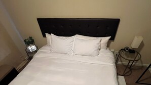 Cama bem arrumada com lençóis brancos, cabeceira escura e mesa de cabeceira com abajur e uma pequena planta.