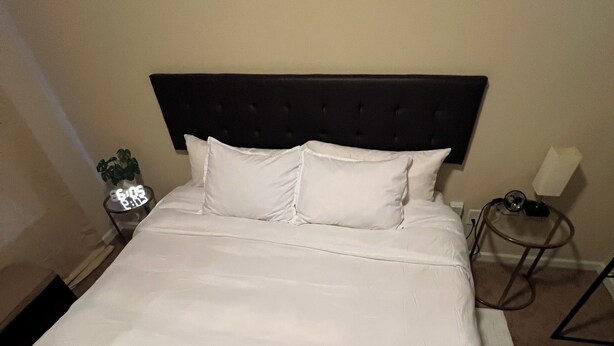 Un lit bien fait, avec du linge de lit blanc, une tête de lit sombre et une table de chevet sur laquelle se trouvent une lampe et une petite plante.