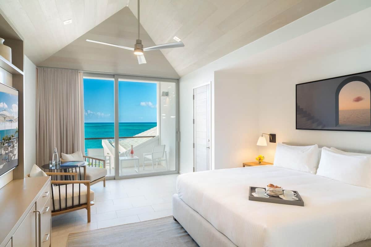 Habitación de hotel con cama, escritorio, silla, balcón con vista al océano y ventilador de techo.