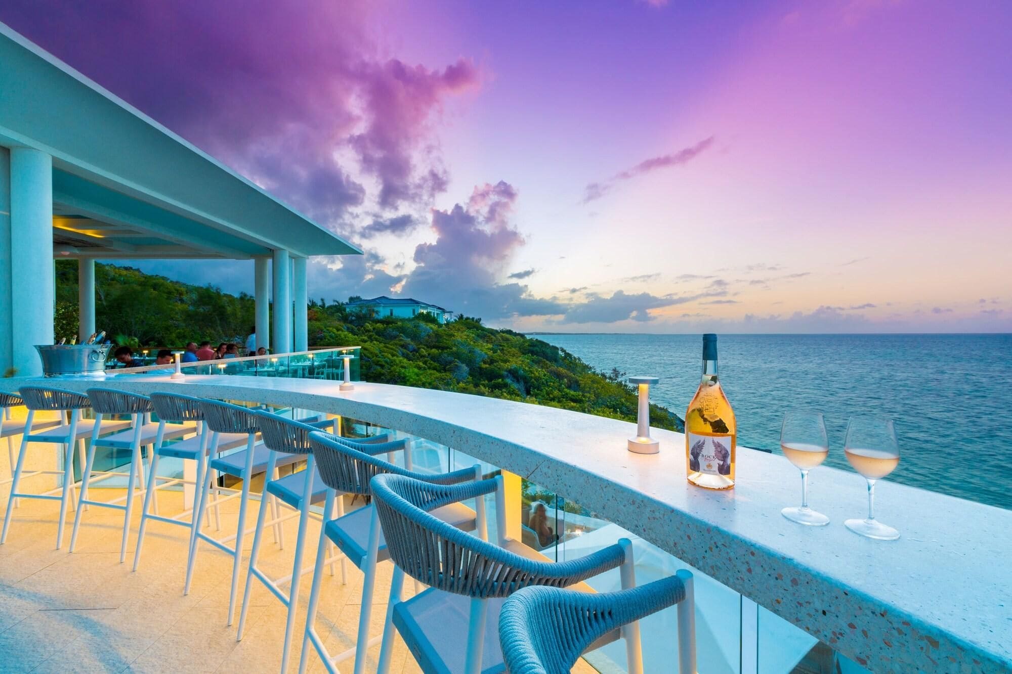 A bar with outdoor seating overlooking the sea, a bottle of wine, and glasses on the counter.