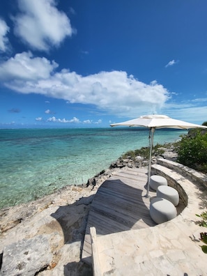 A clear view of a beach with a wooden deck, a white umbrella, and a rocky shoreline. - Rock House (Providenciales)