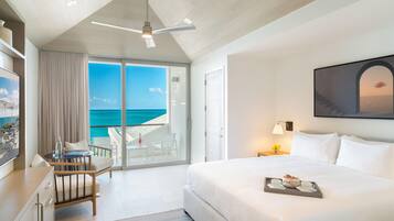 A hotel room with a bed, a desk, a chair, a balcony with a view of the ocean, and a ceiling fan.
