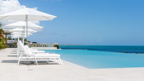 A poolside area with white lounge chairs and umbrellas, a clear blue sky, and a view of the ocean. - Rock House (Providenciales)