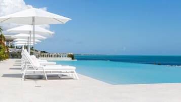 A poolside area with white lounge chairs and umbrellas, a clear blue sky, and a view of the ocean.