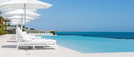Un área junto a la piscina con sillones blancos y sombrillas, un cielo azul despejado y vistas al océano.