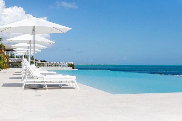 A poolside area with white lounge chairs and umbrellas, a clear blue sky, and a view of the ocean.