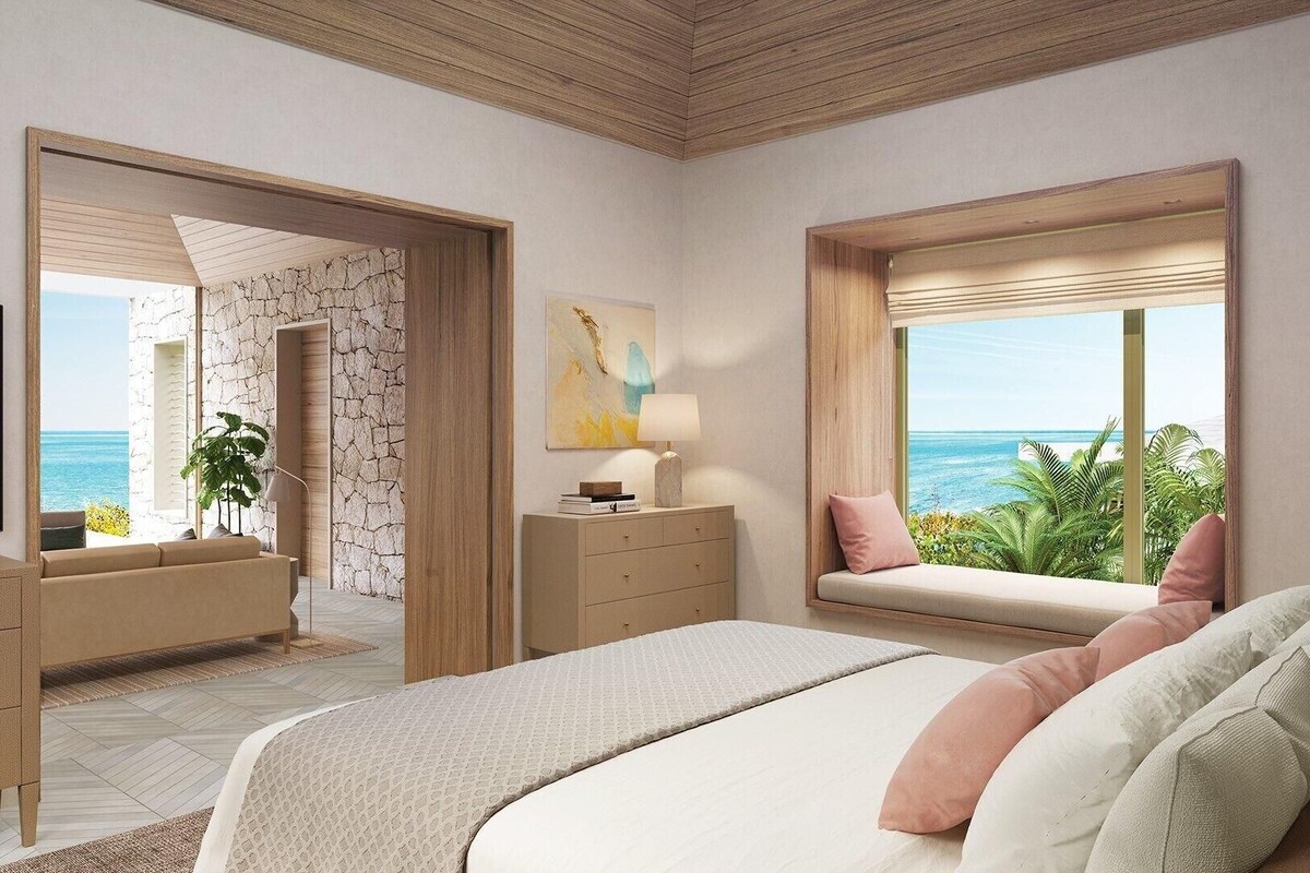 A bedroom with a bed, a dresser, a TV, a window with a view of the ocean, and a plant.