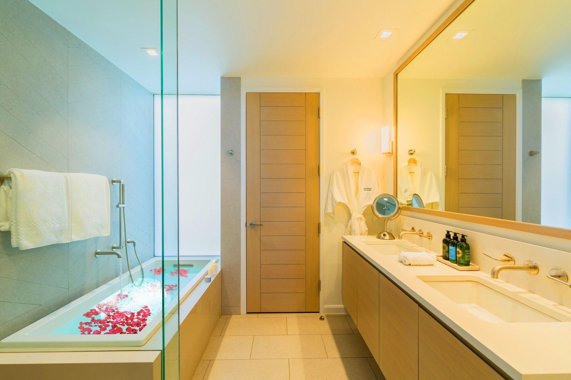 Een moderne badkamer met een douchecabine van glas, een badkuip met rozenblaadjes, een grote spiegel en een wastafel met toiletartikelen.