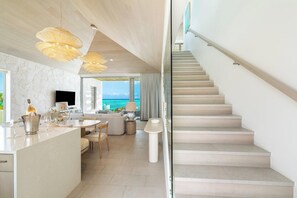 A modern interior with a kitchen, dining area, and a staircase leading to a balcony with a view of the ocean. - Rock House (Providenciales)