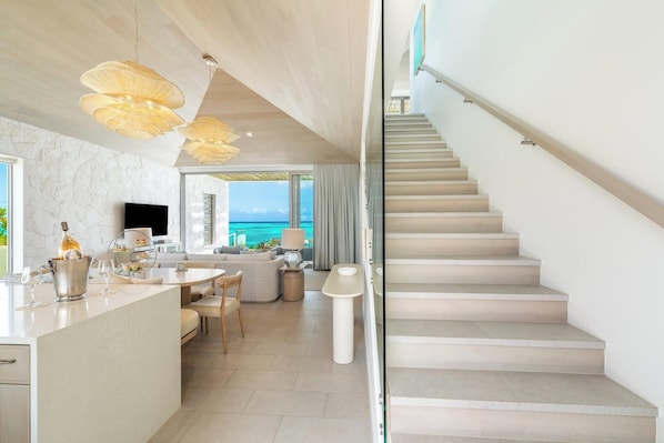 A modern interior with a kitchen, dining area, and a staircase leading to a balcony with a view of the ocean. - Rock House (Providenciales)