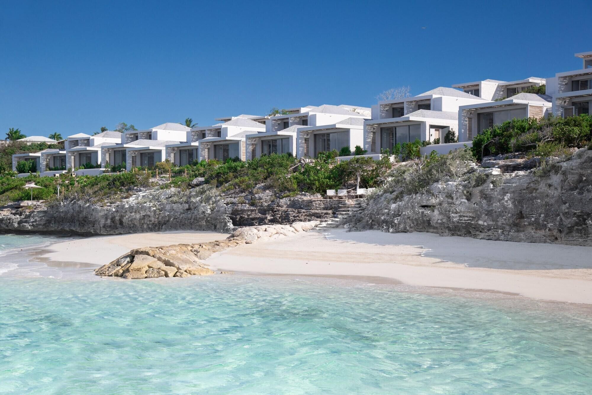 Un hotel frente a la playa con edificios blancos, agua turquesa cristalina y terreno rocoso.