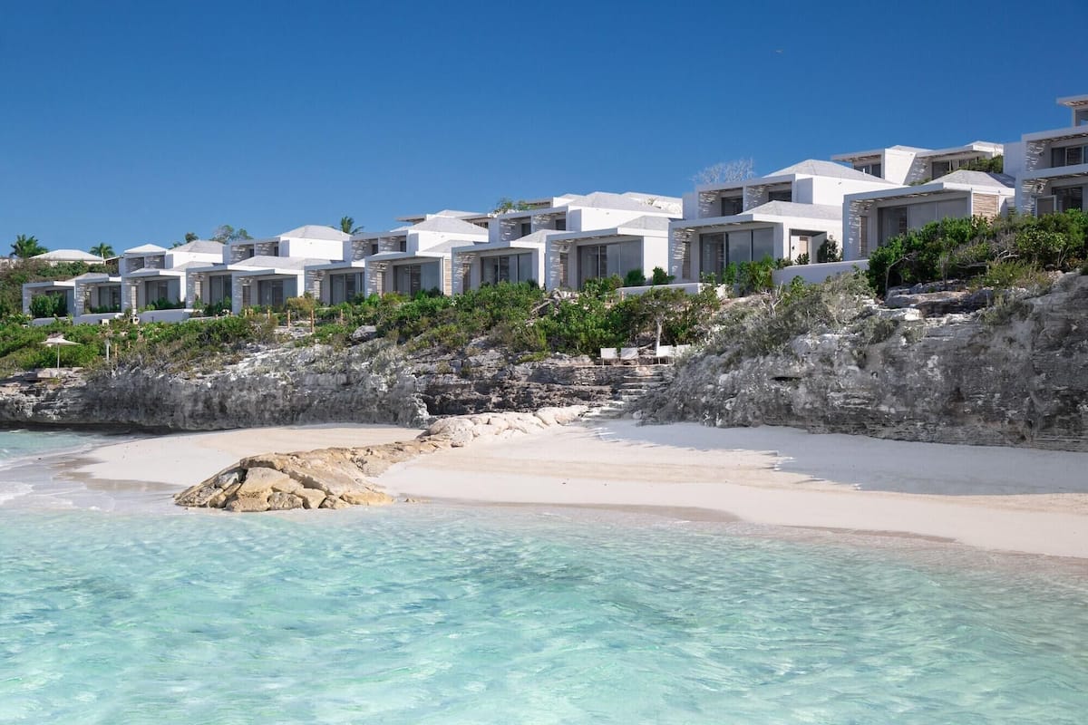 Un hotel frente a la playa con edificios blancos, agua turquesa cristalina y terreno rocoso.