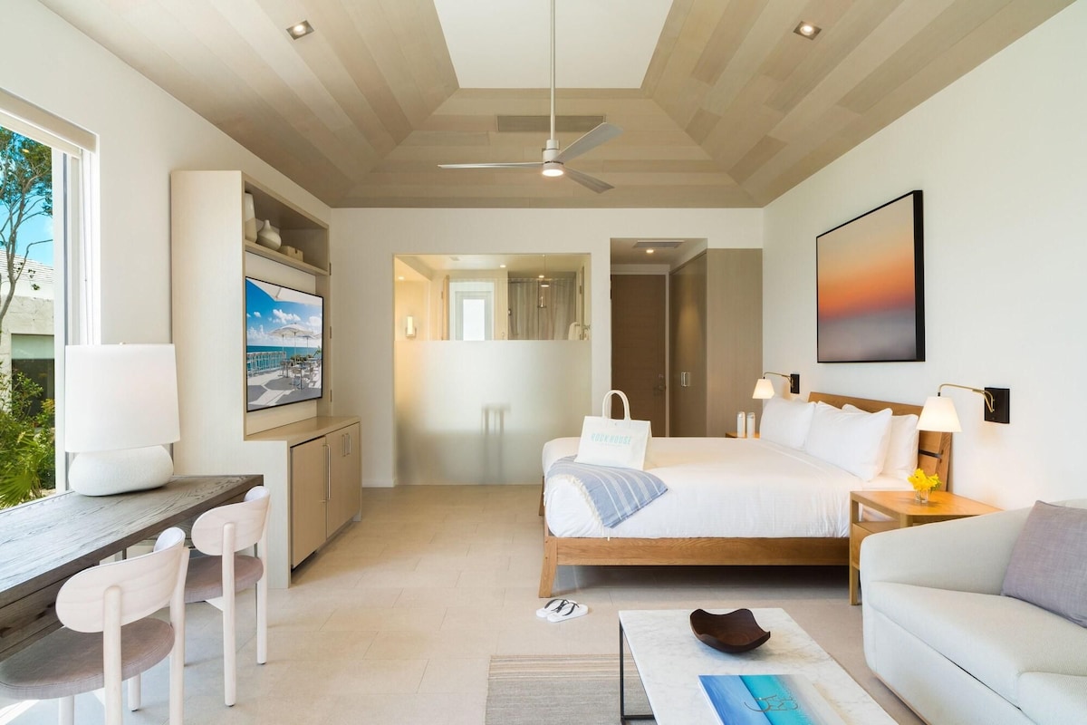 A modern hotel room with a bed, a desk, a chair, a TV, a ceiling fan, and a view of the outdoors.