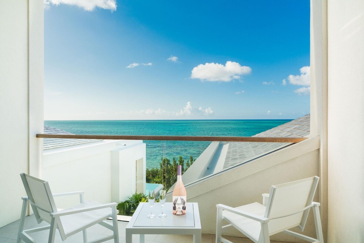 A balcony with a table, two chairs, and a bottle of wine, overlooking a clear blue sea.