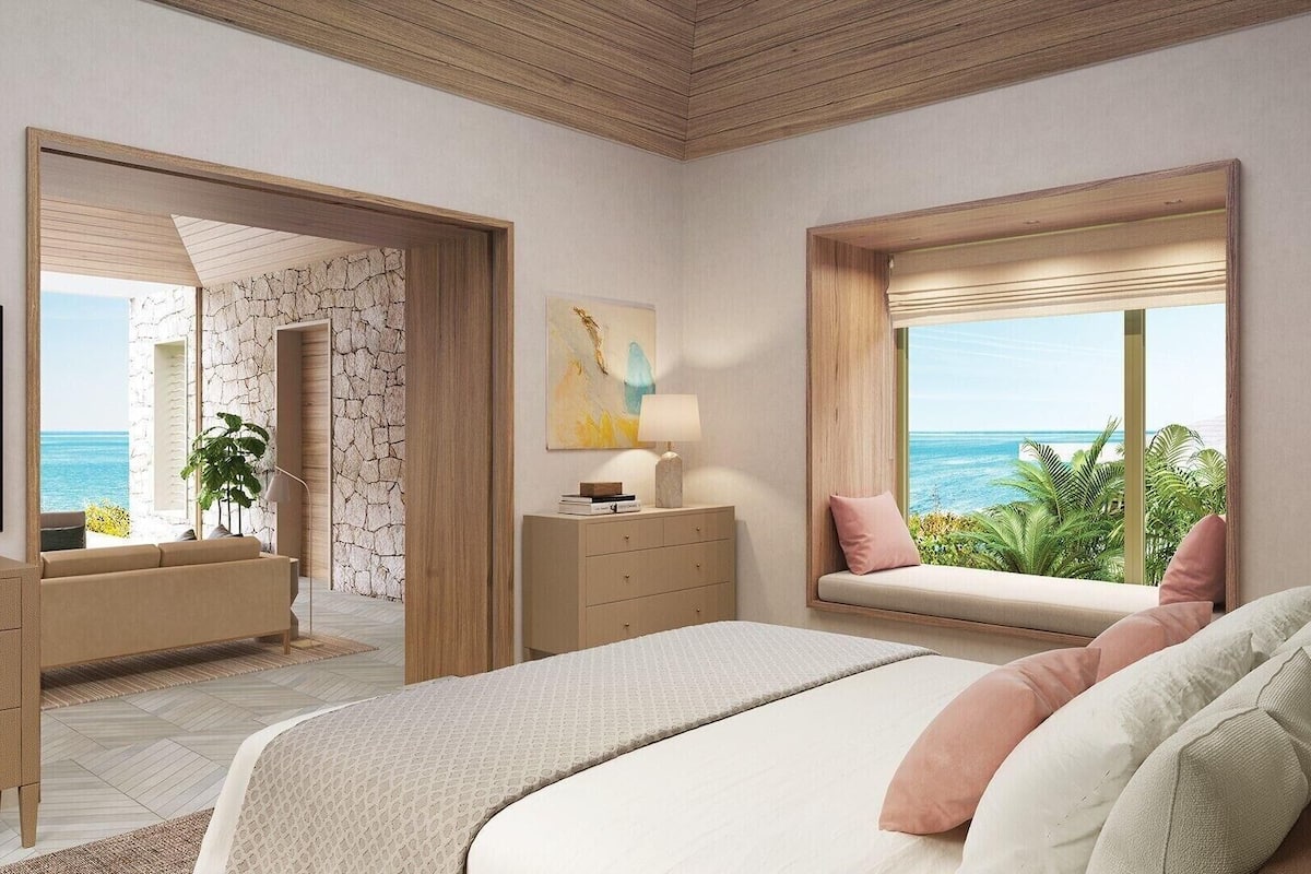 Un dormitorio con cama, cómoda, televisor, ventana y vista al océano, además de una planta.