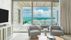 Una habitación moderna con un gran ventanal con vistas a una piscina y al océano, dos sillones, una mesa de centro y un televisor de pantalla plana.