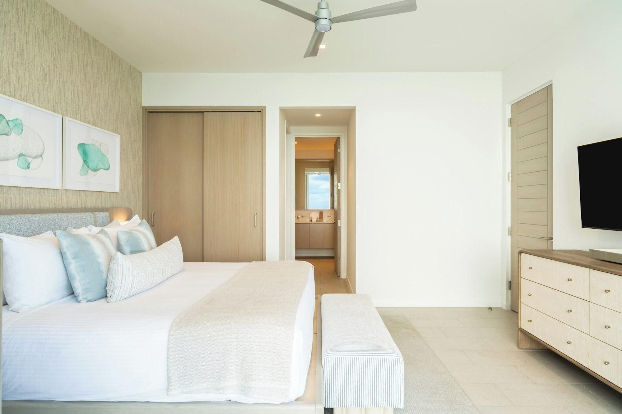 Un dormitorio con cama, un banco, televisor, armario y baño visible a través de una puerta abierta.