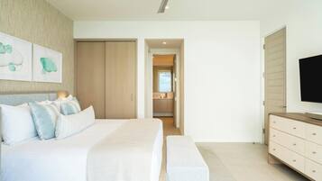 Un dormitorio con cama, un banco, televisor, armario y baño visible a través de una puerta abierta.