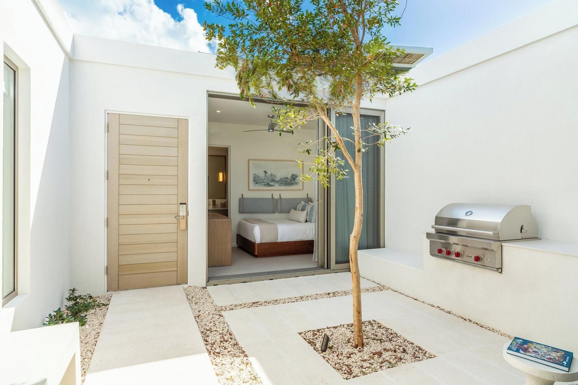 A modern hotel room with a wooden door, a bed, a bathroom, and a barbecue grill.