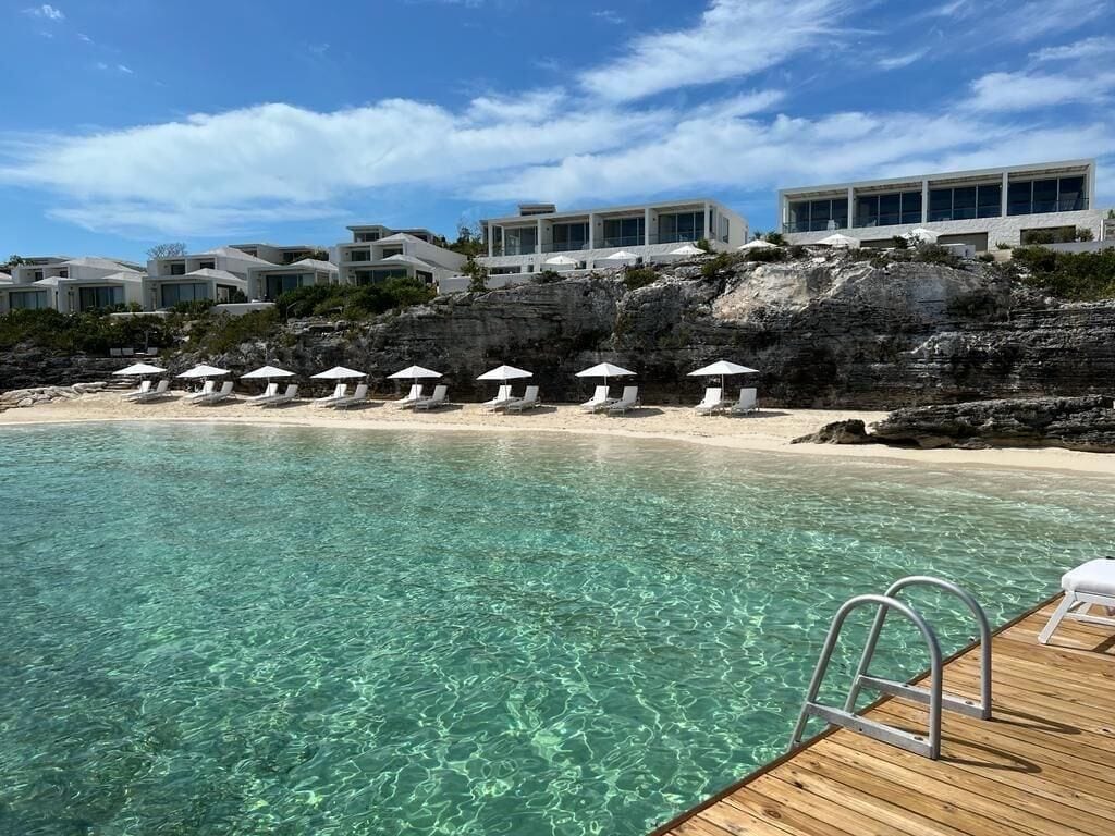 Una piscina cristalina con muelle de madera, sillas de playa y sombrillas, con edificios modernos al fondo.