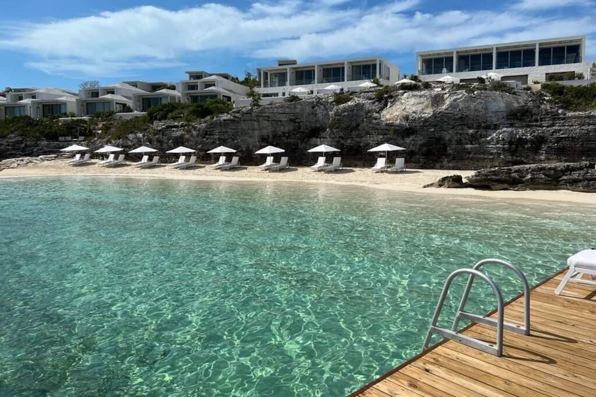 Una piscina cristalina con muelle de madera, sillas de playa y sombrillas, con edificios modernos al fondo.