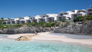 Ett hotell vid stranden med vita byggnader, kristallklart turkosfärgat vatten och en klippig terräng.