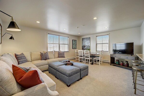 A modern living room with a sectional sofa, ottoman, dining table, chairs, and a flat-screen TV.