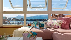 Une pièce confortable avec vue sur un port et des montagnes. Elle comprend un canapé agrémenté de coussins décoratifs, une petite table avec un panier et un saladier de fruits, ainsi qu’une fenêtre donnant sur la mer et les bateaux.