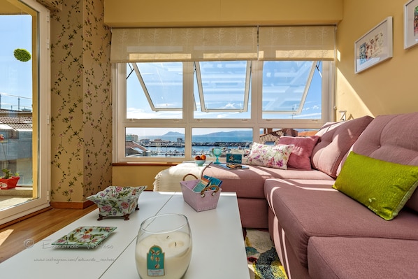 A cozy living room with a pink sofa, a white coffee table, and a view of a harbor through large windows.