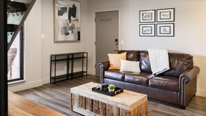 A living room with a brown leather couch, a wooden coffee table, and a black metal console table. There is a framed abstract painting on the wall and a door in the background. - Silvertown by AvantStay | Gorgeous Home in Fantastic Location in Park City! (Park City)