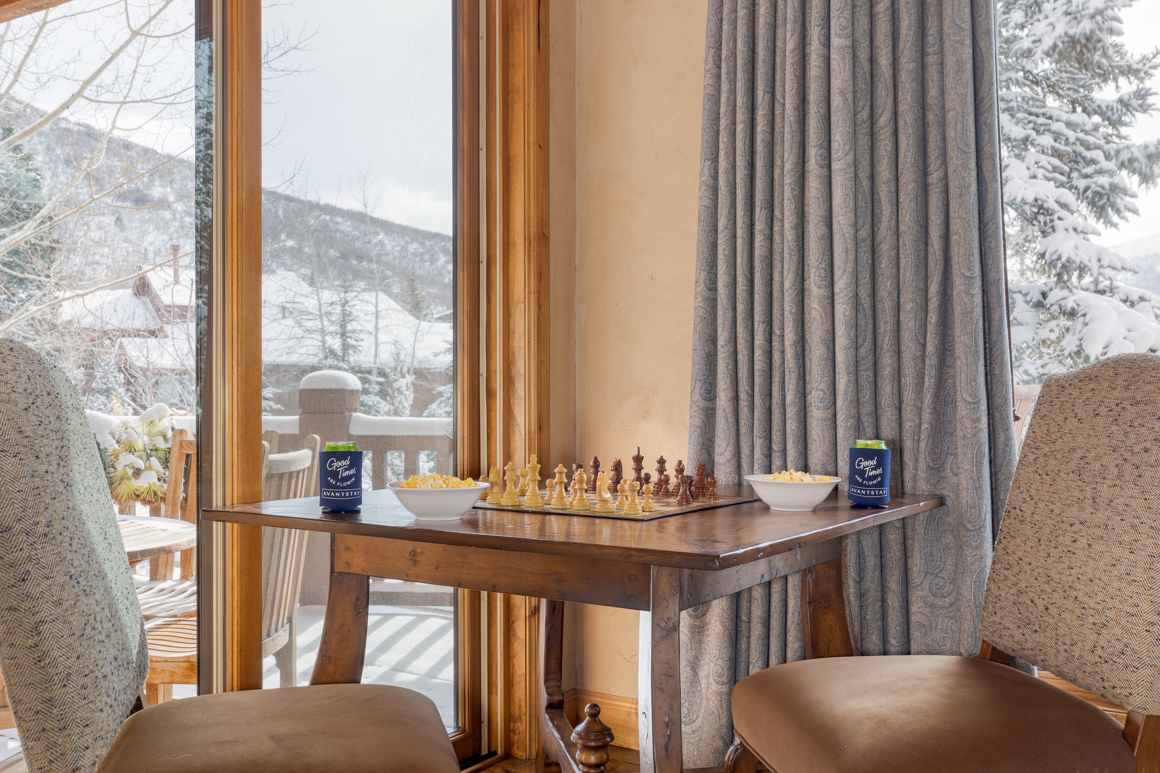 A cozy room with a chessboard on a table, a bowl of snacks, and a view of snow-covered mountains.