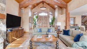 A spacious living room with a wooden ceiling, a stone wall, a large flat-screen TV, a wooden coffee table, and a beige sofa with blue cushions.
